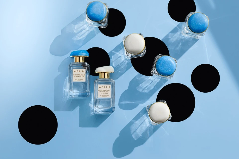 Two AERIN perfume bottles and several solid perfume compacts scattered on a light blue background, with black geometric shapes adding contrast.
