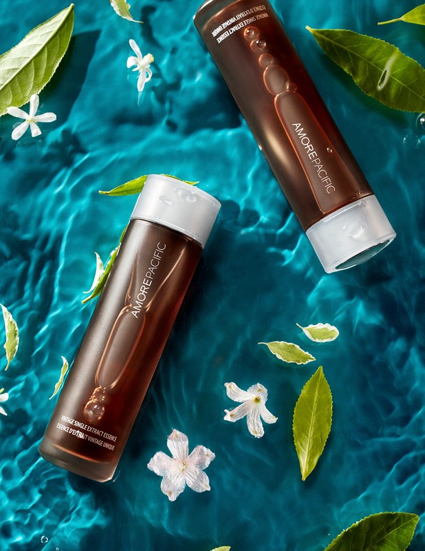 Two brown AmorePacific bottles on a blue water-like surface with white flowers and green leaves create a fresh, beauty product display.