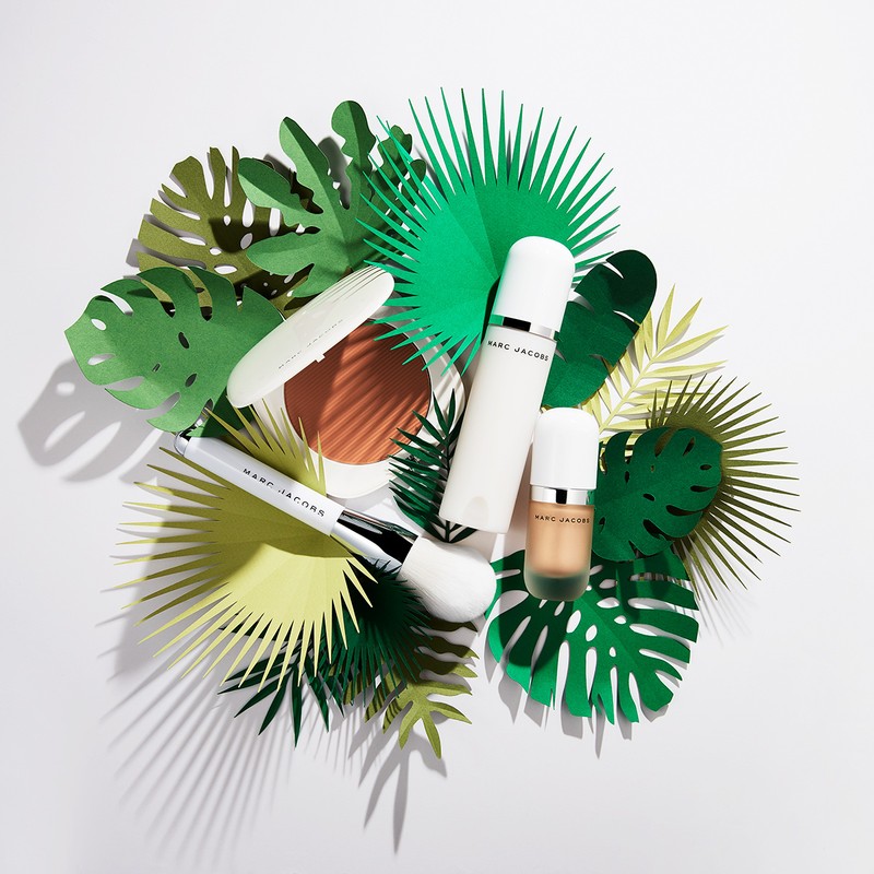 Marc Jacobs makeup products including foundation, powder, and brush, surrounded by green paper palm leaves, against a white backdrop.