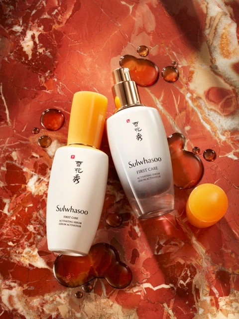 Sulwhasoo skincare product bottles in white and gold on a red marble surface with amber liquid droplets, product photography.