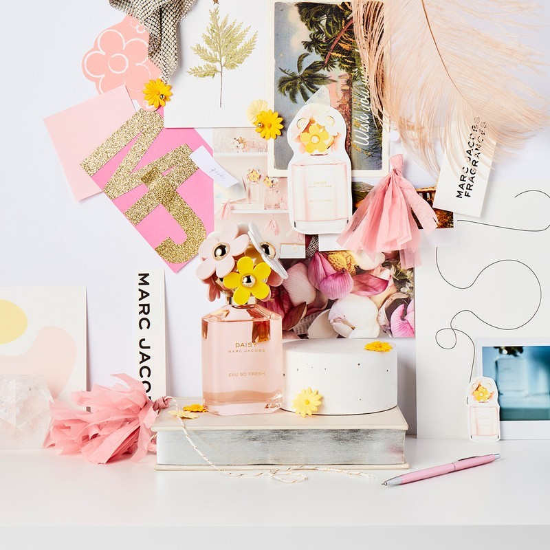 Still life studio shot of Marc Jacobs Daisy Eau So Fresh perfume bottle, placed on a stack of books, surrounded by floral and paper decorations.