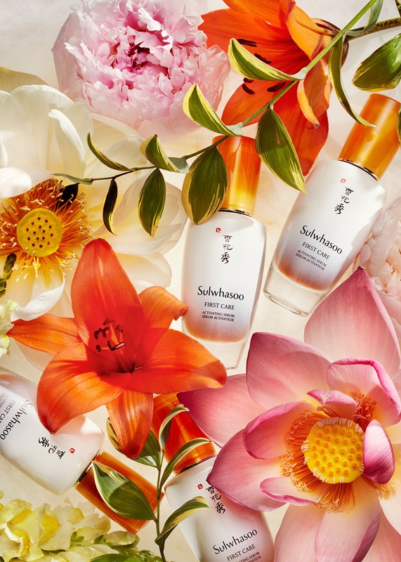 Floral arrangement featuring Sulwhasoo First Care serum bottles nestled among vibrant lilies, peonies, and lotuses against a bright background.