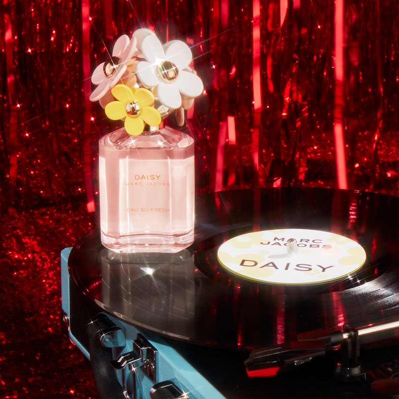 Marc Jacobs Daisy Eau So Fresh perfume bottle on a record player with red glitter background, emphasizing floral design and brand.