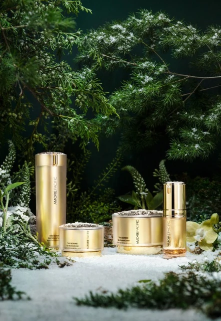 Golden skincare product containers are arranged among evergreen branches dusted with snow against a dark green backdrop in a studio shot.