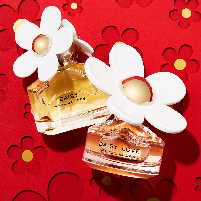 Two bottles of Marc Jacobs Daisy perfume on a red background with daisy cutouts, showcasing a luxury fragrance product shot.
