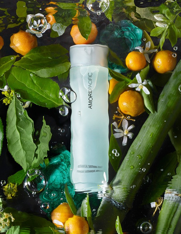 Product shot of AmorePacific Botanical Soothing Toner in a clear bottle, surrounded by citrus fruit, leaves, and flowers in a water setting.