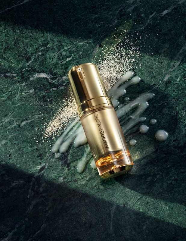 Gold Amorepacific serum bottle on green marble with scattered powder and liquid for beauty and skincare advertising, product photography.
