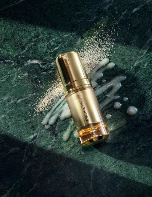 Gold Amorepacific serum bottle on green marble with scattered powder and liquid for beauty and skincare advertising, product photography.