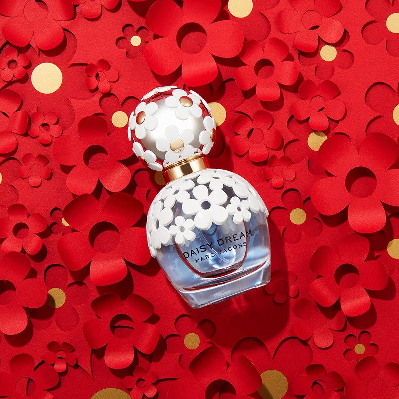 Daisy Dream perfume bottle by Marc Jacobs on a bed of red paper flowers and gold confetti. Cosmetic product shot. Beauty aesthetic.