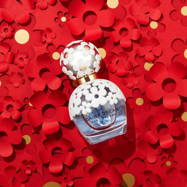 Daisy Dream perfume bottle by Marc Jacobs on a bed of red paper flowers and gold confetti. Cosmetic product shot. Beauty aesthetic.