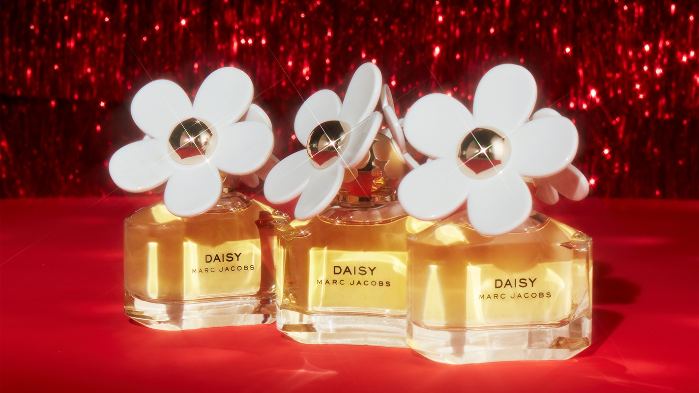 Three Marc Jacobs Daisy perfume bottles with white floral caps on a red surface, set against a sparkling red background.