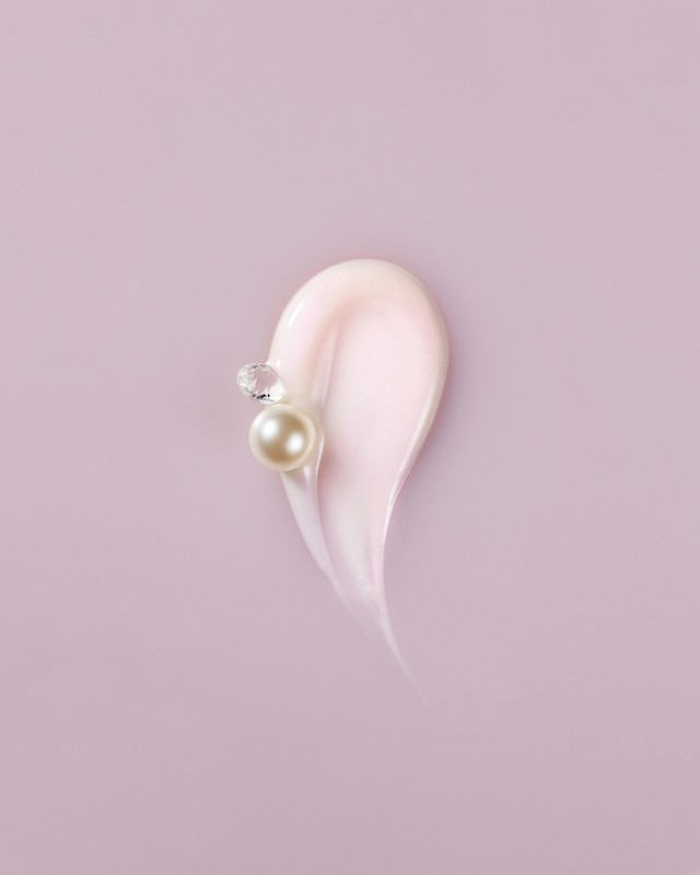 Close-up of light pink cosmetic cream smear on pale purple background with white pearl and clear gem. Beauty and skincare concept.