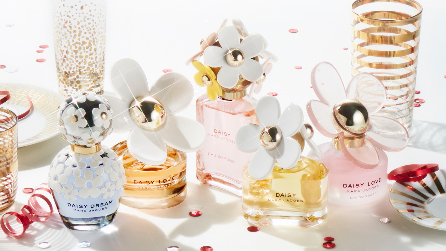 Close-up studio shot of multiple Marc Jacobs Daisy perfume bottles with daisy-themed caps, confetti, and decorative glassware.
