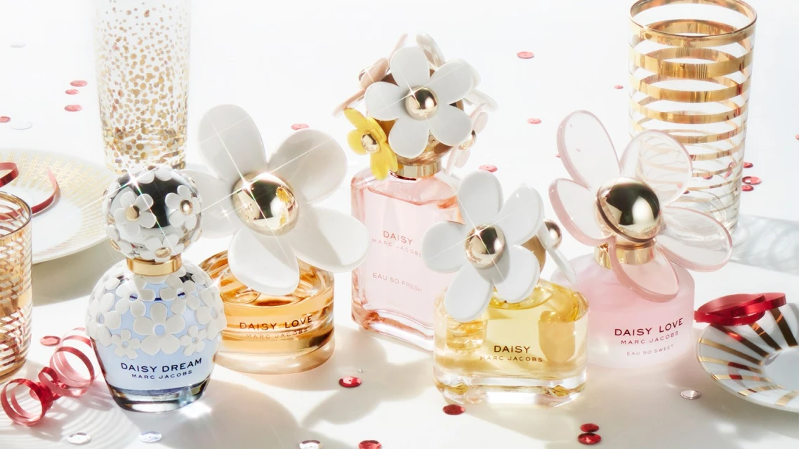 Close-up studio shot of multiple Marc Jacobs Daisy perfume bottles with daisy-themed caps, confetti, and decorative glassware.