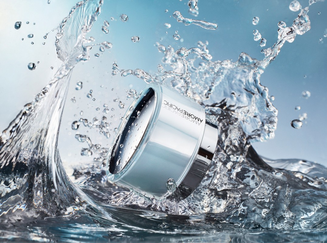 AmorePacific cream jar surrounded by splashing water against a gradient blue background for a beauty product advertisement.