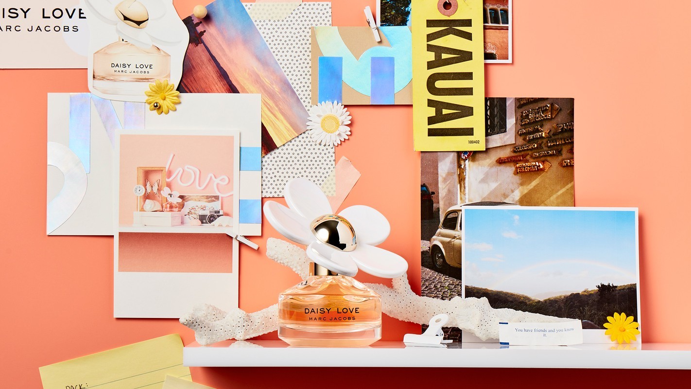 Daisy Love Marc Jacobs perfume bottle surrounded by cheerful paper ephemera and warm coral backdrop, evoking a lighthearted and youthful vibe.