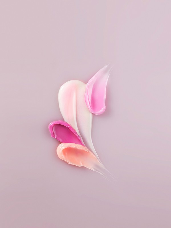 Various shades of lip gloss or lipstick are smeared on a pastel background, featuring pink, peach, and cream tones in vertical patterns.