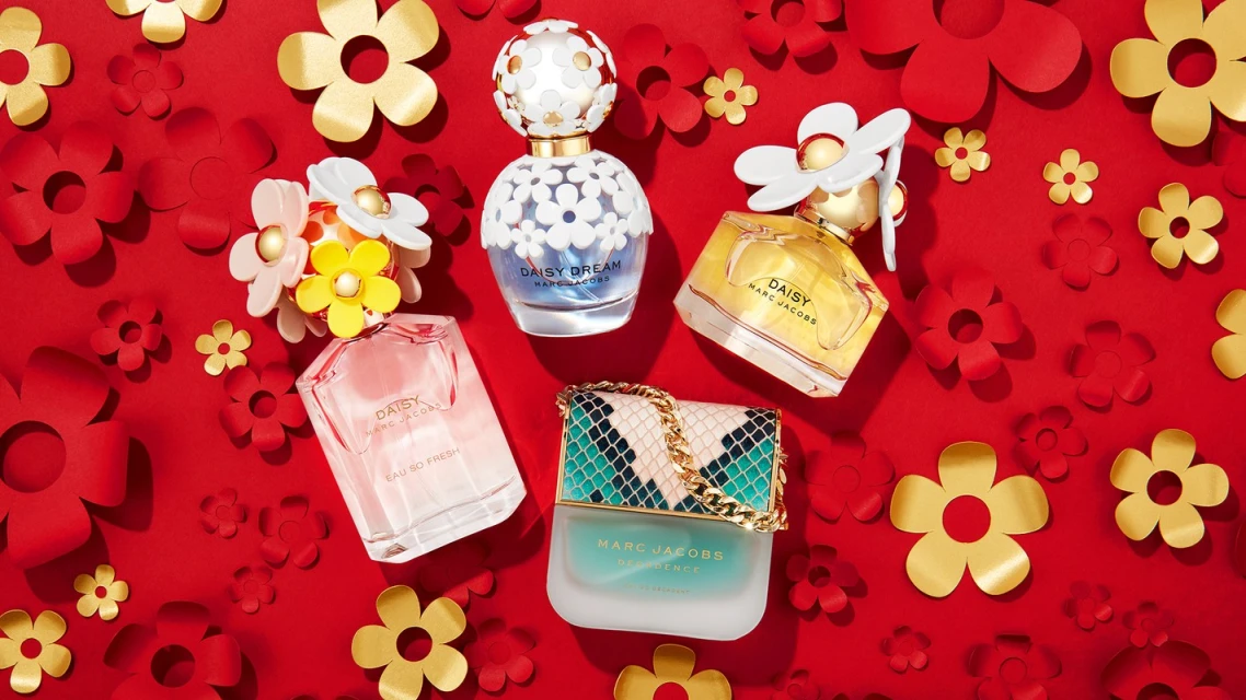Five bottles of Marc Jacobs Daisy fragrances are arranged on a vibrant red background adorned with decorative gold and red paper flowers.