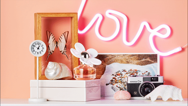 Still life composition features a perfume bottle, shells, a clock, framed butterfly, and a neon light against a peach-colored backdrop.