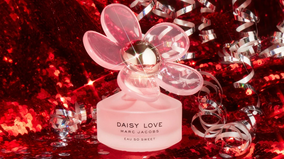Pink flower-shaped Daisy Love perfume bottle by Marc Jacobs, placed on a red sequined surface with silver streamers. Beauty product.