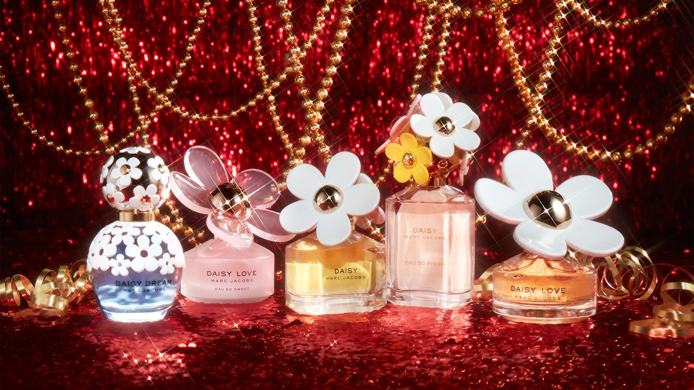 Five Marc Jacobs Daisy perfume bottles on a red glitter surface, adorned with gold chains and ribbons, captured in a studio shot.