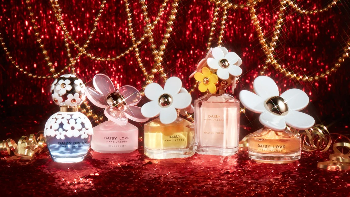 Five Marc Jacobs Daisy perfume bottles on a red glitter surface, adorned with gold chains and ribbons, captured in a studio shot.