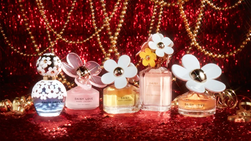 Five Marc Jacobs Daisy perfume bottles on a red glitter surface, adorned with gold chains and ribbons, captured in a studio shot.