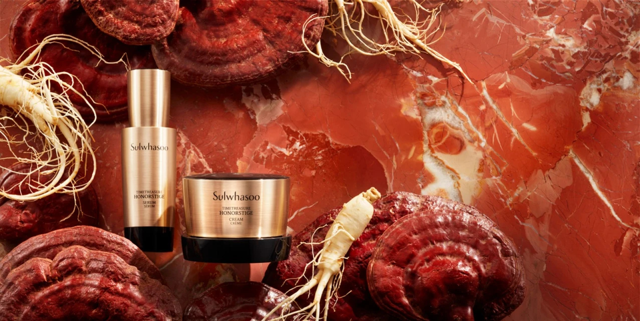 Two Sulwhasoo TimeTreasure Honorstige cream and serum products nestled amongst red reishi mushrooms and ginseng roots against red marble.