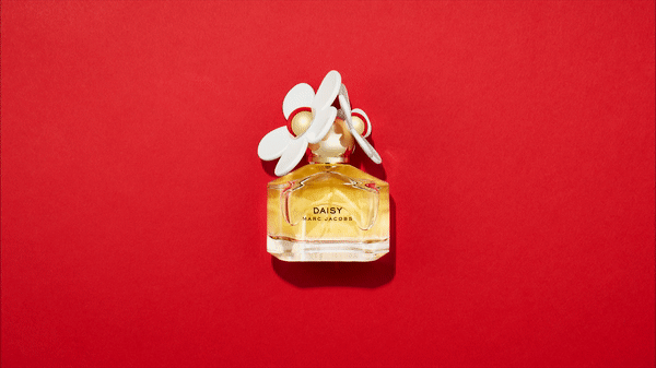 Top-down studio shot of a perfume bottle with a daisy cap on a red background. The bottle is half-filled with gold liquid.