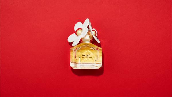 Top-down studio shot of a perfume bottle with a daisy cap on a red background. The bottle is half-filled with gold liquid.