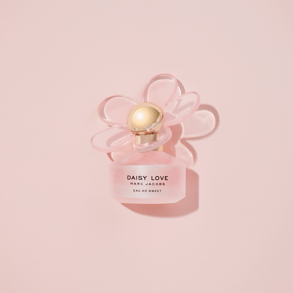 Close-up studio shot of a 'Daisy Love Marc Jacobs' perfume bottle with daisy-shaped cap on a light pink backdrop, product photography.