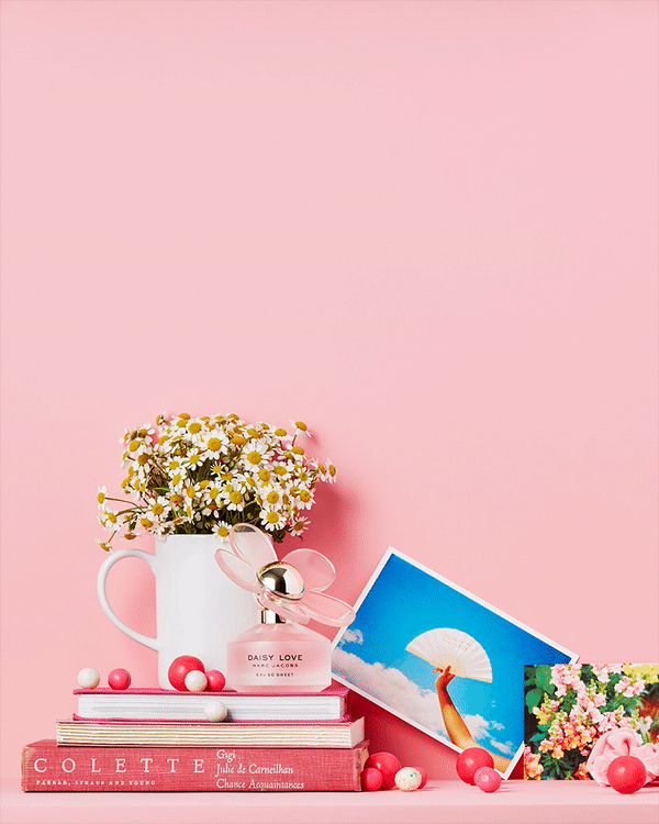 Daisy perfume bottle displayed on a stack of vintage books with floral arrangements and pink background, commercial beauty shot.
