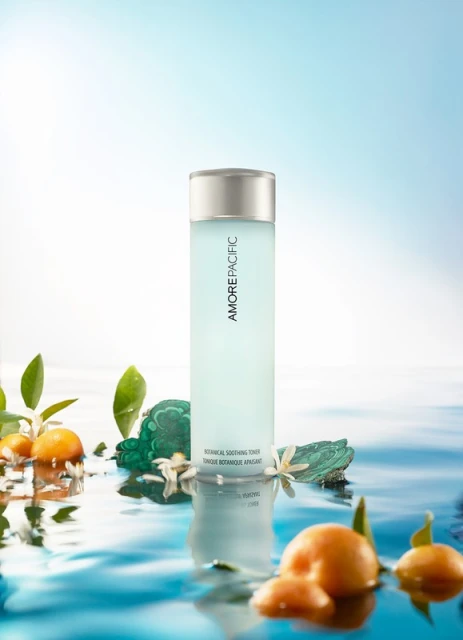 Bottle of AMOREPACIFIC skincare tonic surrounded by oranges, green stones, and white flowers in a studio setting with blue and white background.