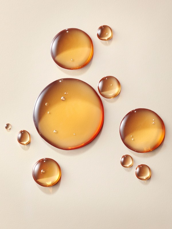 Close-up shot of various sized drops of translucent, amber-colored liquid on a light tan surface creating an abstract, textured cosmetic background.