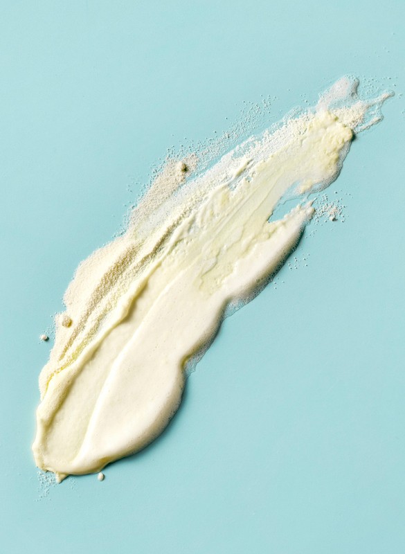 Swath of yellowish-white cosmetic cream or lotion applied across a light blue background in a studio-lit, top-down skincare product shot.