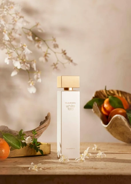 Elizabeth Arden White Tea perfume bottle sits on a wooden surface with tangerines and white flowers in a soft, warm-toned studio shot.