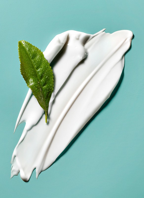 Close-up of white cream swipe on light blue surface with green leaf. Beauty product shot for skincare or natural cosmetics ad.