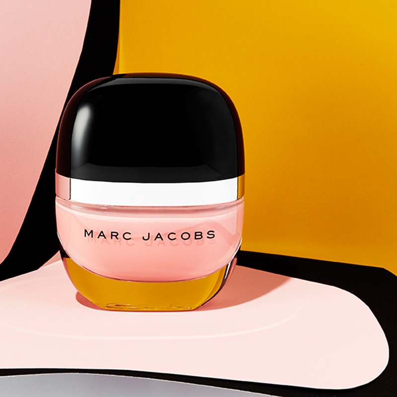 Close-up studio shot of a Marc Jacobs beauty product in a black and silver container, with a pink tinted substance, against a geometric backdrop.