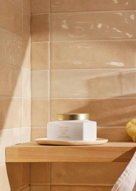 A square jar of Elizabeth Arden White Tea body cream sits on a wood shelf in a bathroom, near a tan tiled wall.