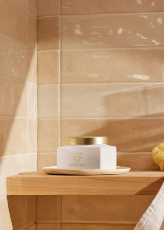 A square jar of Elizabeth Arden White Tea body cream sits on a wood shelf in a bathroom, near a tan tiled wall.