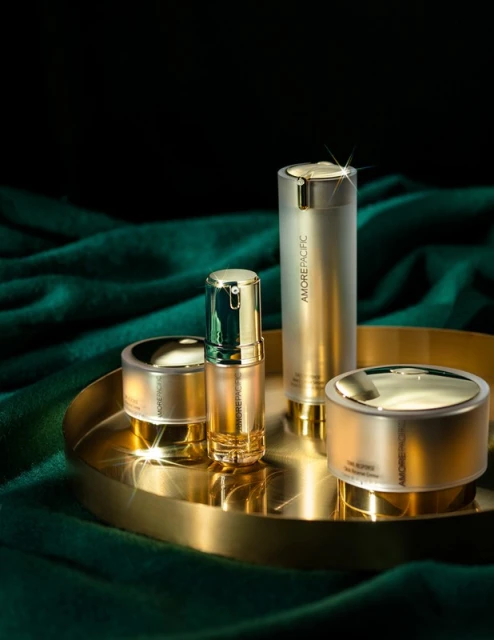 Four golden Amorepacific skincare products arranged on a golden tray with a dark green velvet draped background, creating a luxurious beauty still life.