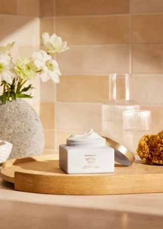 Cosmetic cream jar on a wooden tray with flowers, glass bottles, and a sponge in a brightly lit beige-toned indoor setting.