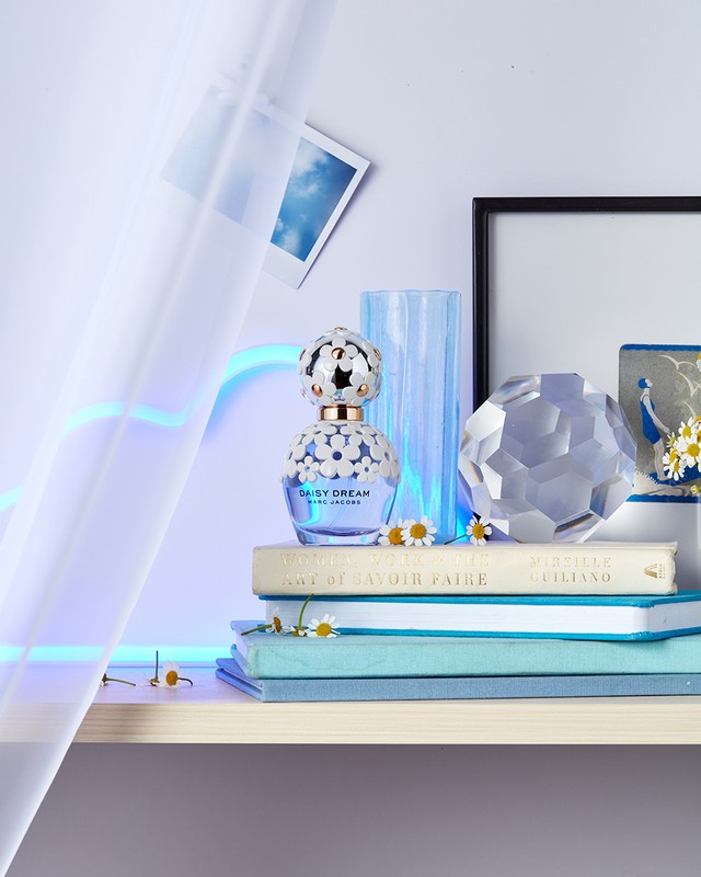 Daisy Dream perfume bottle by Marc Jacobs on books with small daisies, soft light, and a white sheer curtain in the background.