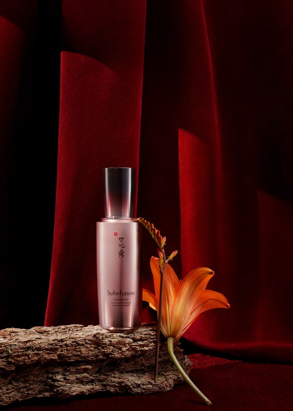 Still life shot of a Sulwhasoo serum bottle placed on a piece of wood with an orange lily against a dark red velvet backdrop.