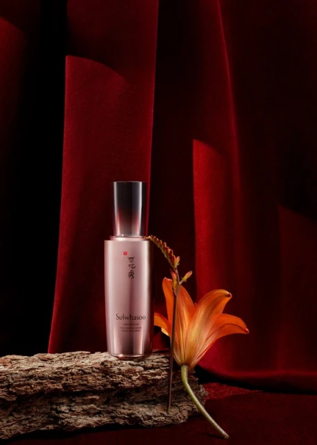 Still life shot of a Sulwhasoo serum bottle placed on a piece of wood with an orange lily against a dark red velvet backdrop.