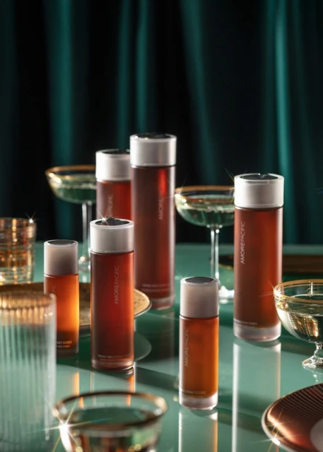 Group of Amorepacific skincare bottles arranged with champagne glasses on a reflective surface, set against a draped fabric background.