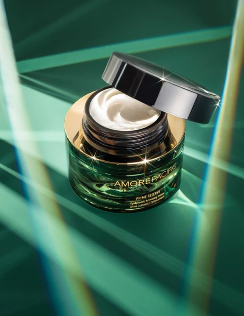 Close-up of AmorePacific cream jar and lid on a green surface with light refraction patterns, emphasizing skincare and product presentation.