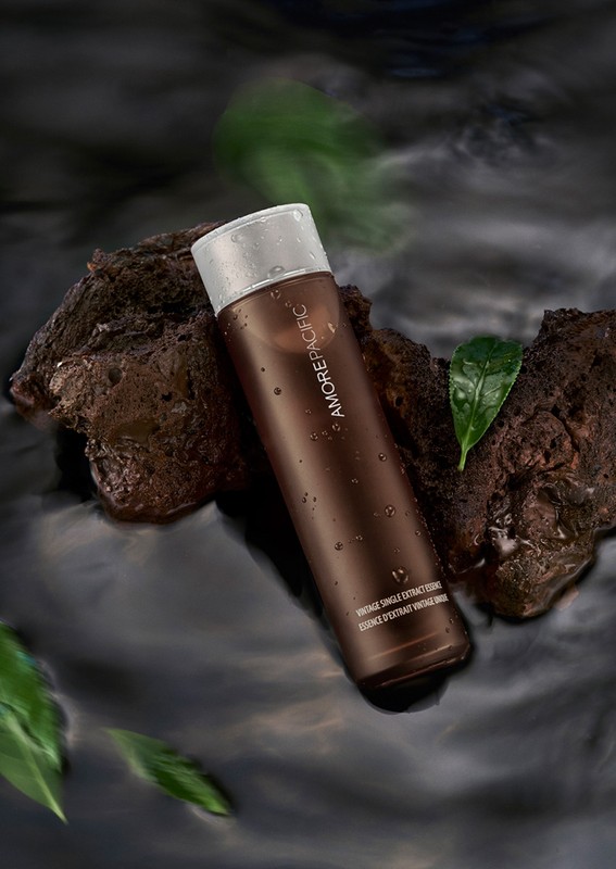 Brown bottle of Amorepacific skincare product resting on a rock in water with green leaves, set against a dark background.