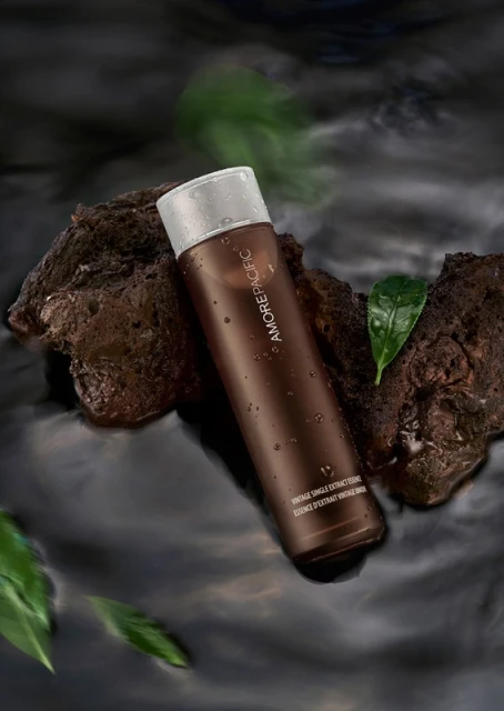 Brown bottle of Amorepacific skincare product resting on a rock in water with green leaves, set against a dark background.