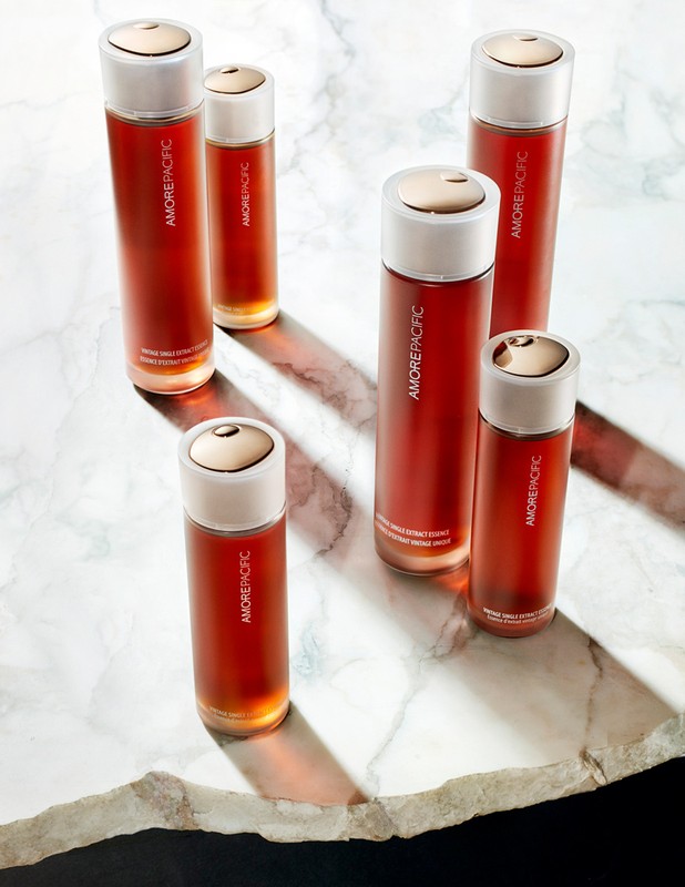 Six amber-colored glass skincare bottles with white and gold caps are arranged on a white marble surface, illuminated by strong side lighting.